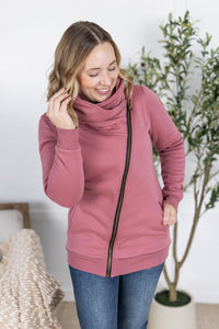Woman wearing a pink zip-up sweatshirt in a home setting with a plant in the background