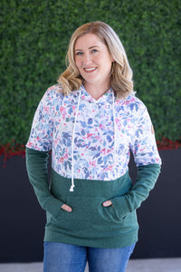 Woman wearing a floral hoodie and green pants standing against a green hedge.