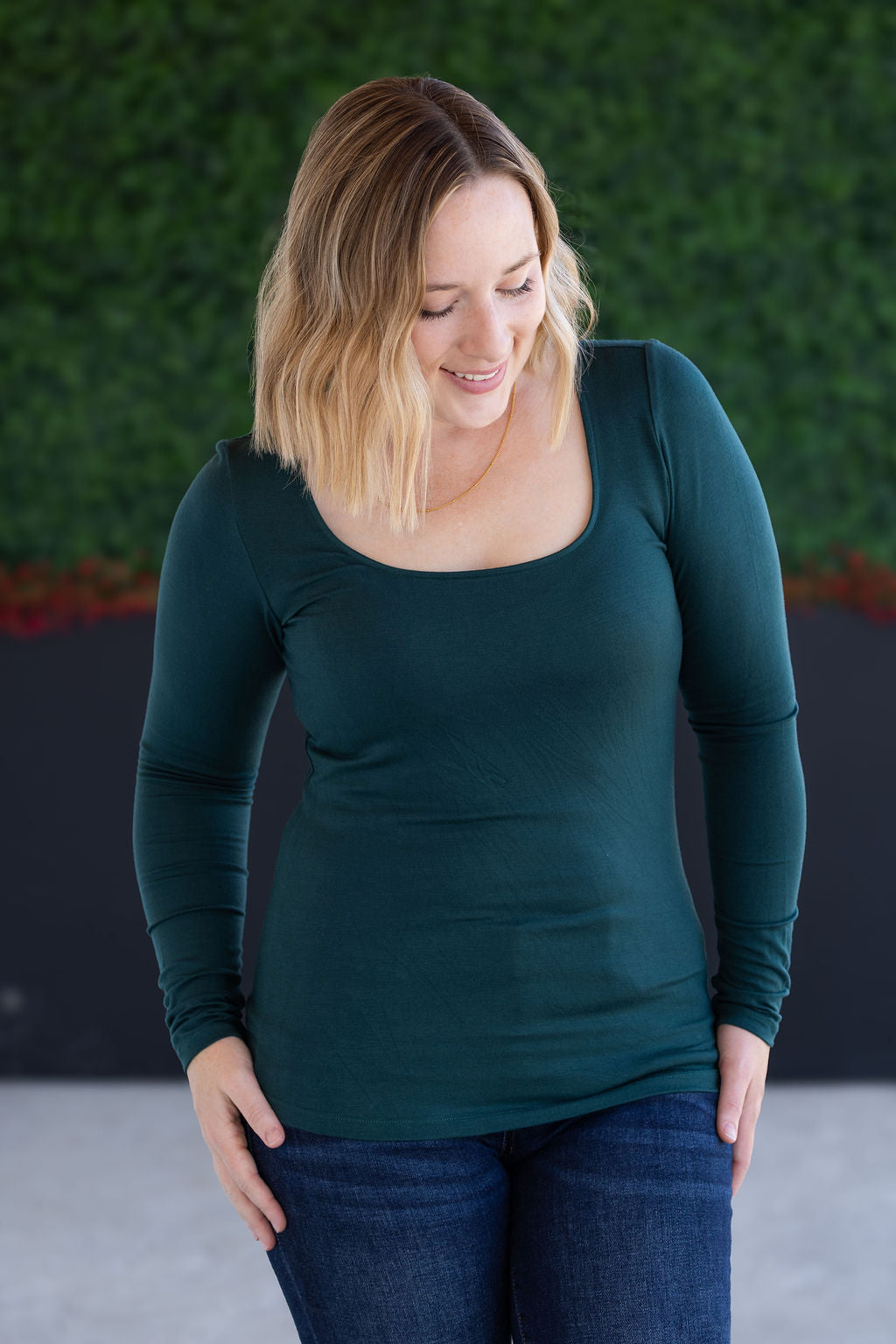 Woman wearing a green long-sleeve top and blue jeans against a green wall.