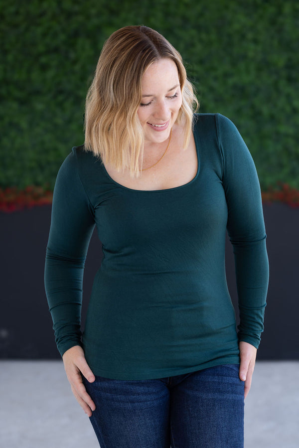 Woman wearing a green long-sleeve top and blue jeans against a green wall.