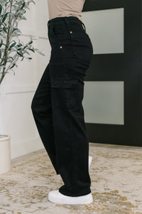 Person wearing black cargo pants standing indoors with a plant and abstract art in the background.