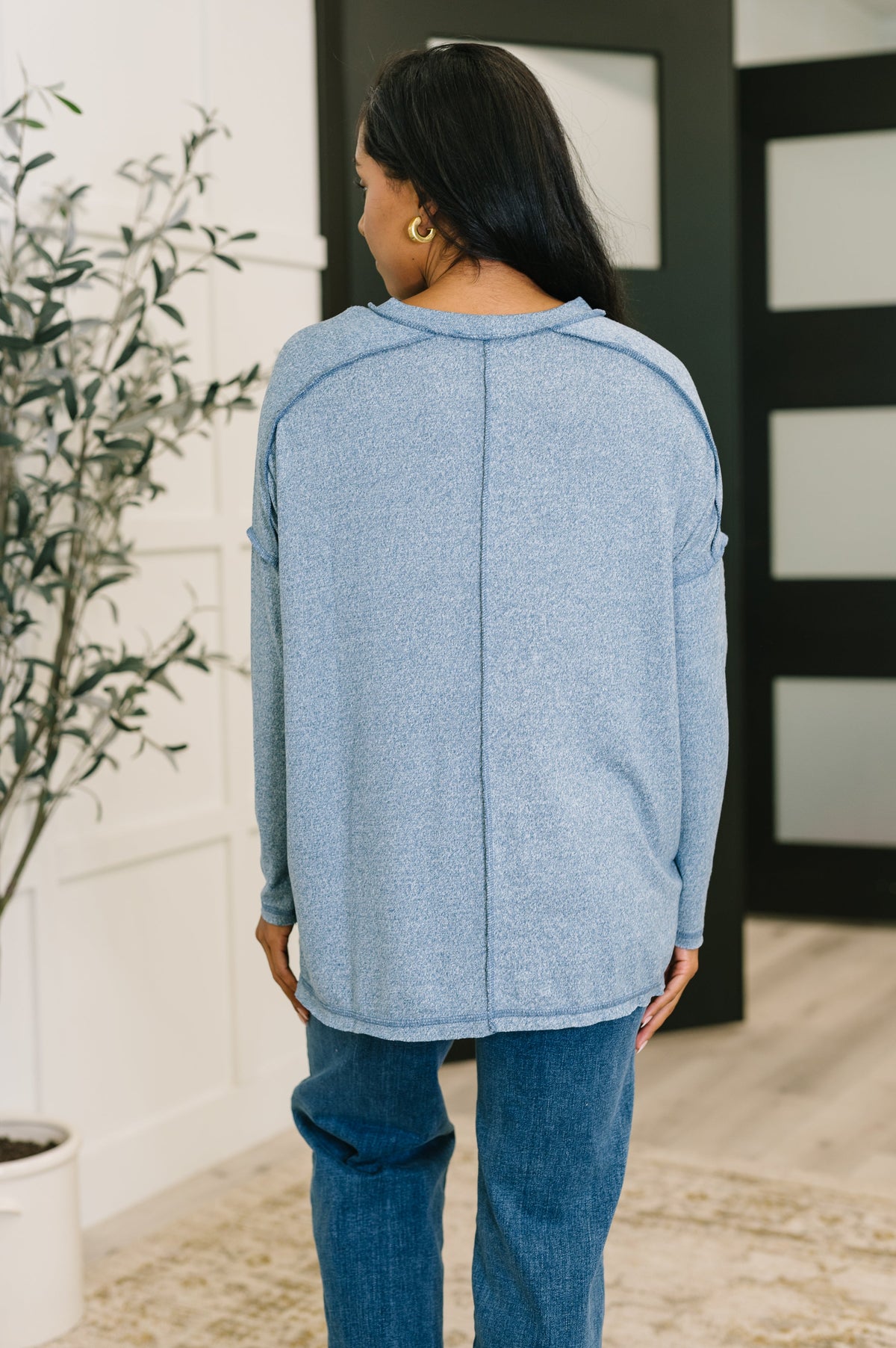 Person wearing a blue sweater and jeans in an indoor setting with a plant and wall decor.