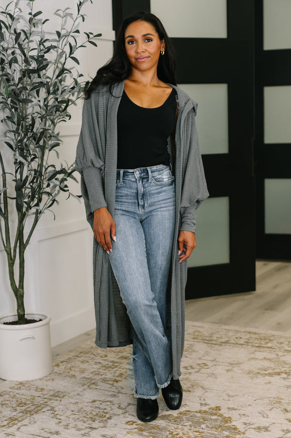 Woman wearing a long gray cardigan, black top, and blue jeans standing indoors.