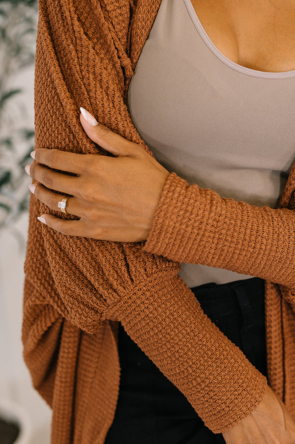 Person wearing a brown knitted cardigan over a beige top with a blurred background