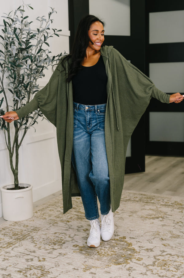 Woman wearing a green cardigan, black top, and blue jeans indoors.