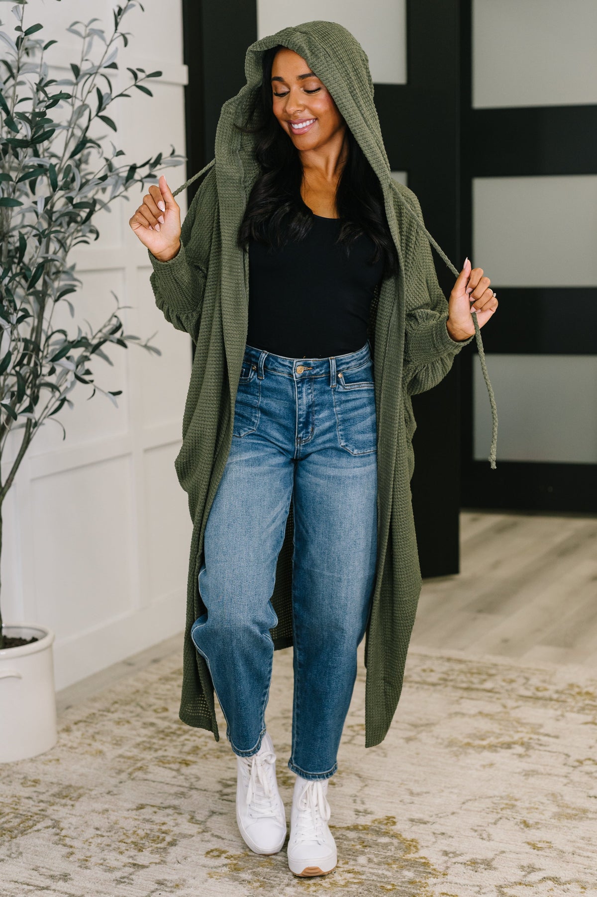 Woman wearing a long green cardigan, black top, and blue jeans indoors.