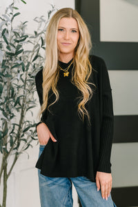 Woman wearing a black sweater and blue jeans standing indoors with a plant in the background