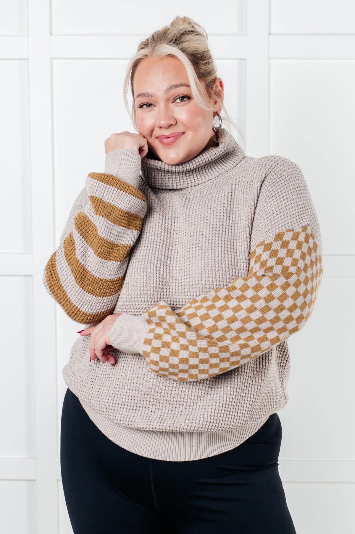 Woman wearing a beige sweater with patterned sleeves against a white tiled wall.