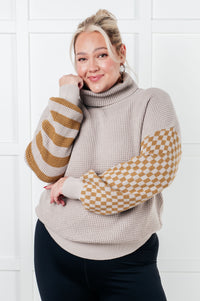 Woman wearing a beige sweater with patterned sleeves against a white tiled wall.