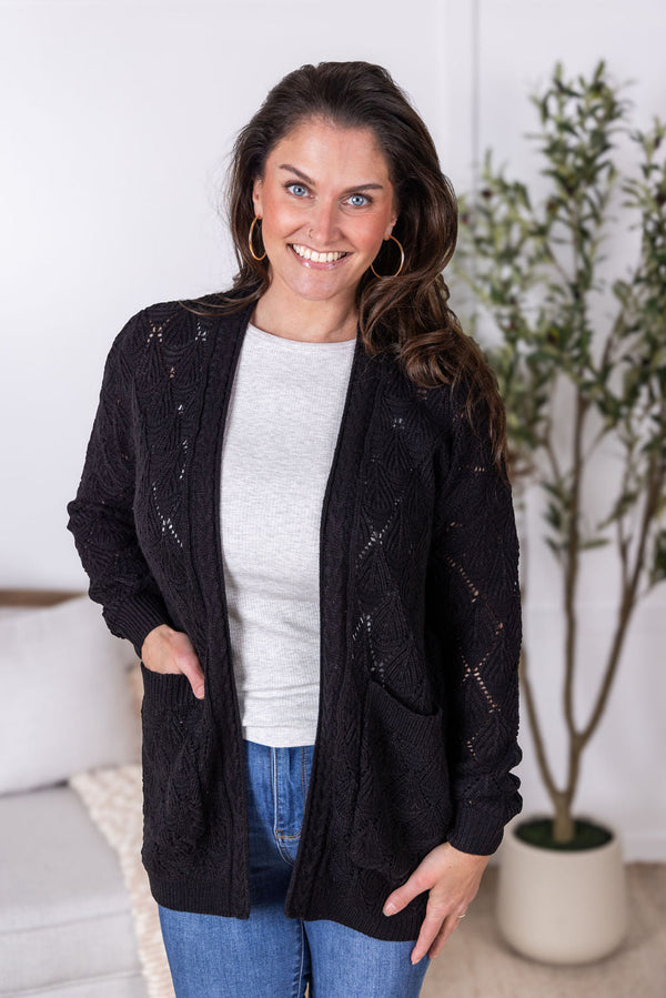 Woman wearing a black cardigan over a white shirt and blue jeans, standing indoors.