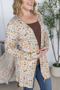 Woman wearing a floral cardigan over a brown top and blue jeans, standing indoors.