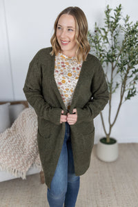Woman wearing a green cardigan over a floral shirt and blue jeans, standing indoors with a plant in the background.