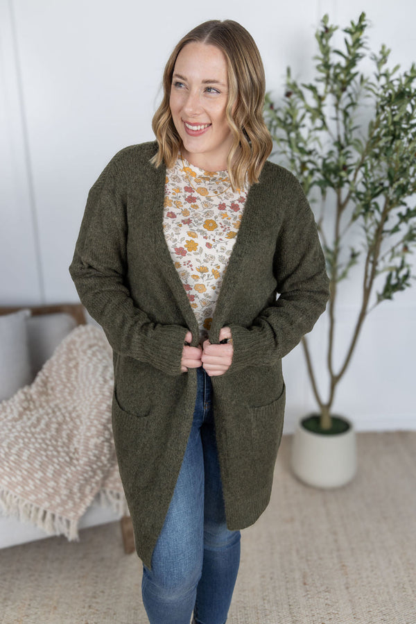 Woman wearing a green cardigan over a floral shirt and blue jeans, standing indoors with a plant in the background.