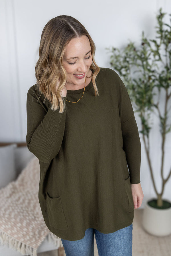 Woman wearing an olive green sweater in a home setting with a plant in the background