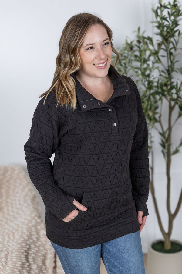 Woman wearing a black quilted jacket with a white wall and plant in the background