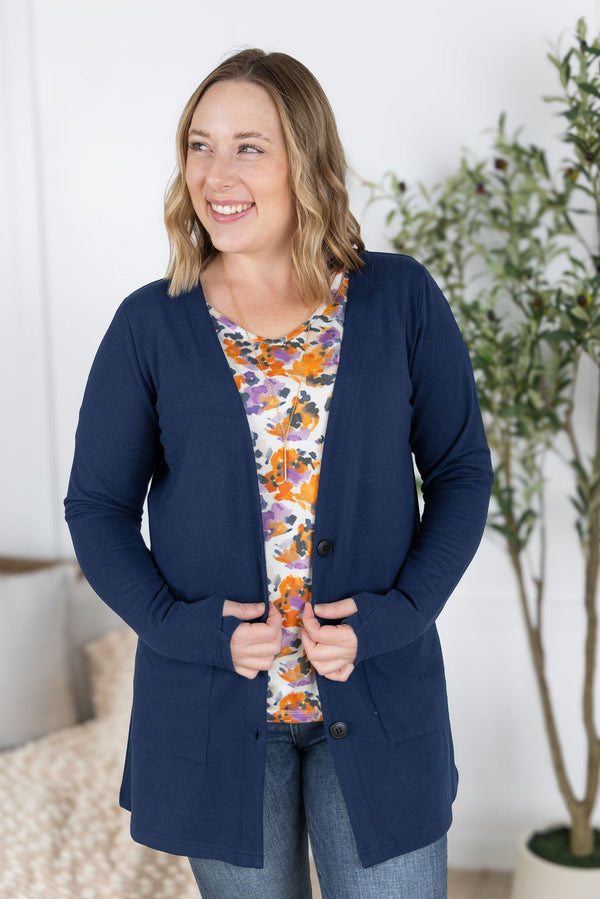 Woman wearing a navy cardigan over a floral top in a home setting with a plant in the background.