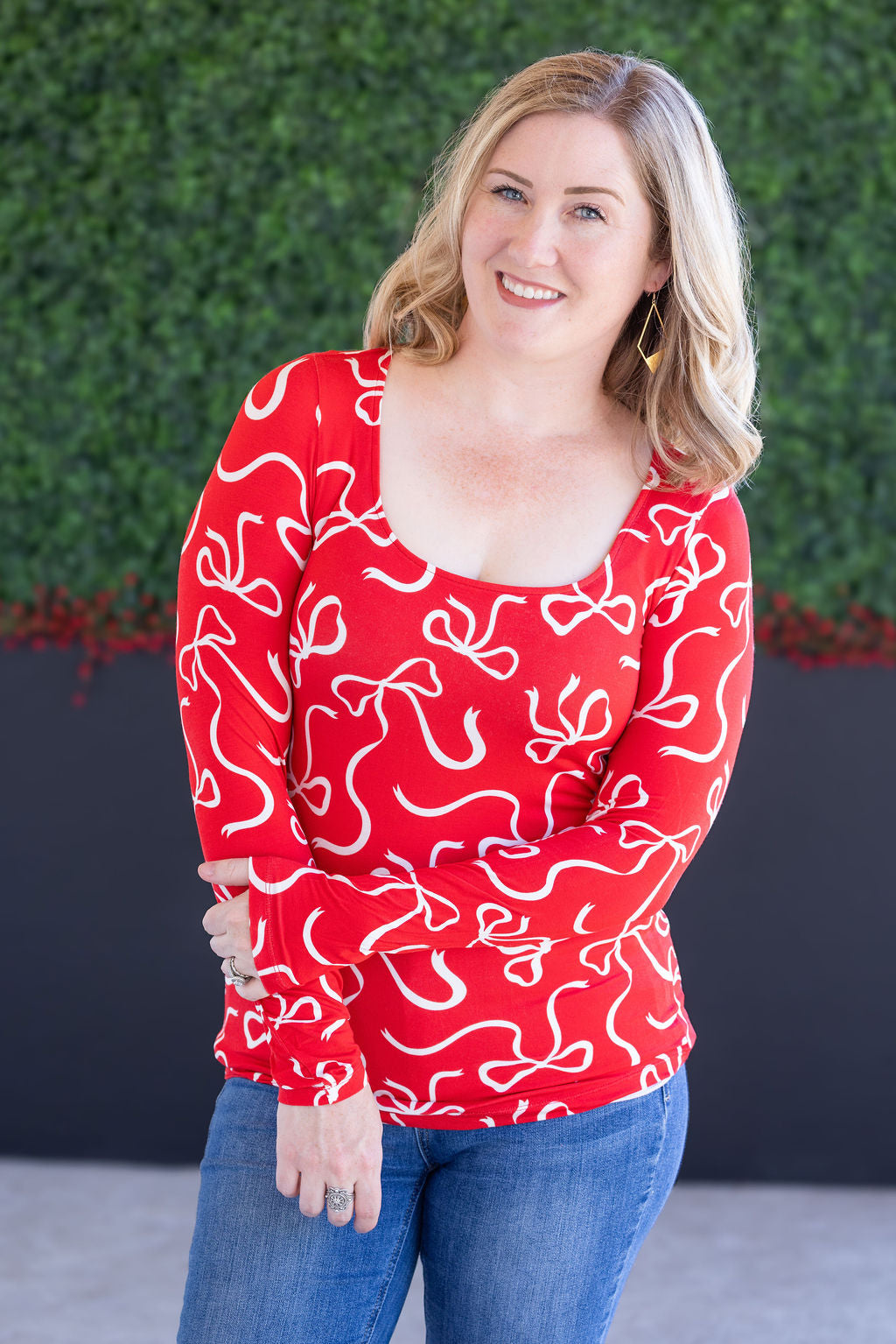 Woman wearing a red patterned top with blue jeans against a green hedge background