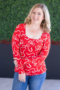 Woman wearing a red patterned top with blue jeans against a green hedge background