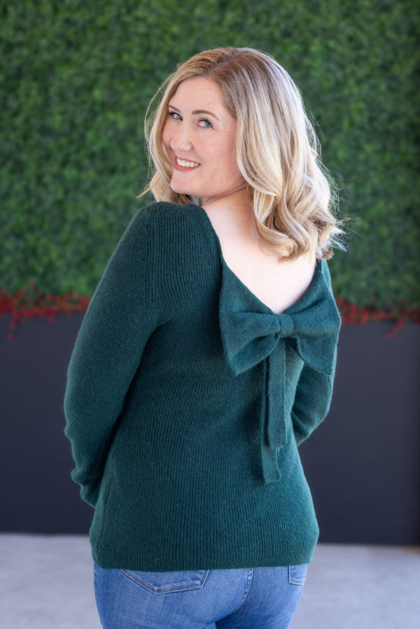 Woman wearing a green sweater with a bow detail, standing against a green wall.