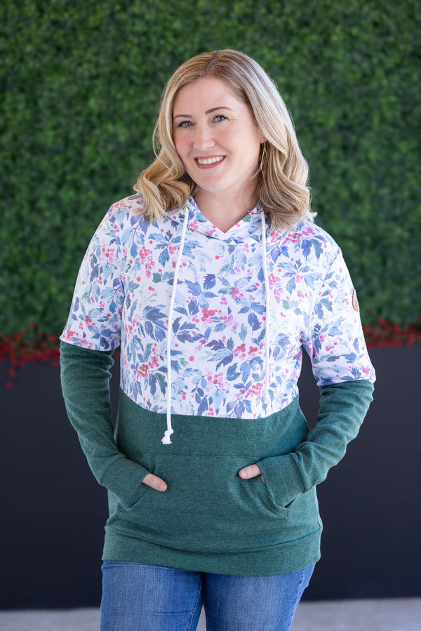 Woman wearing a floral hoodie and green pants standing against a green hedge.