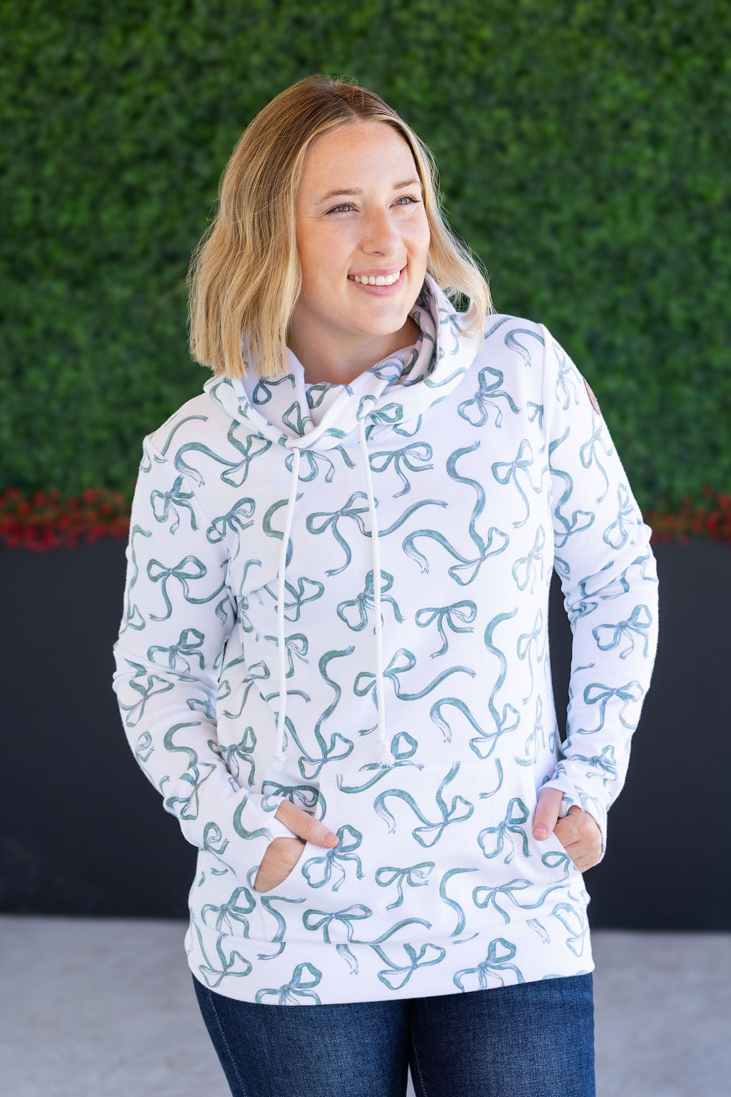 Woman wearing a white hoodie with blue patterns against a green hedge background