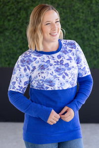 Woman wearing a blue and white floral sweater with a solid blue bottom half against a green hedge background.