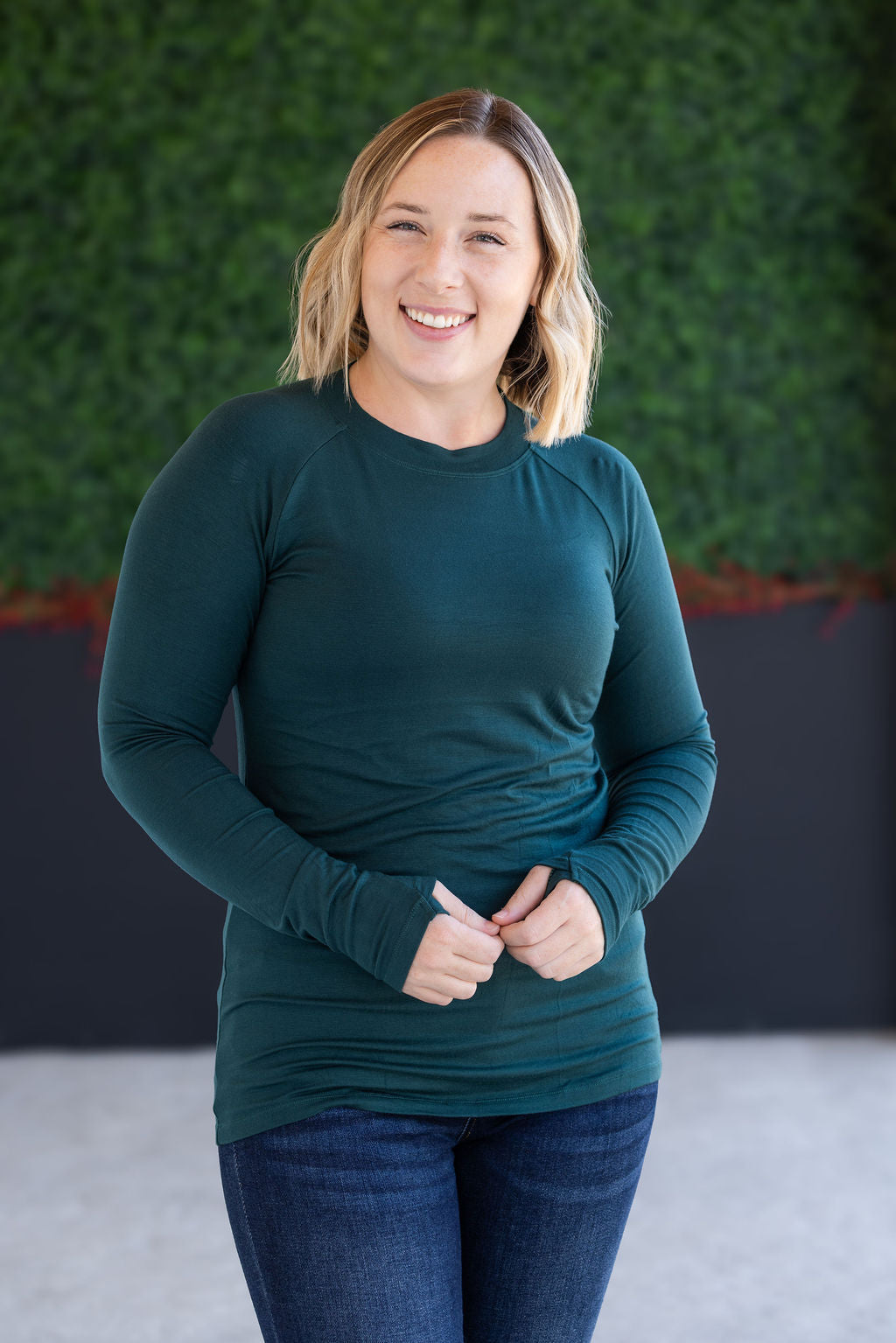 Woman wearing a teal long-sleeve top and blue jeans against a green wall.