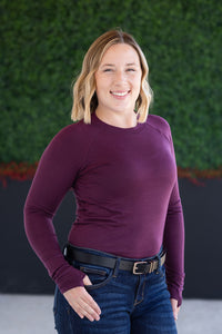 Woman wearing a purple long-sleeve top and blue jeans against a green wall.