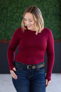 Woman wearing a red long-sleeve top and blue jeans against a green wall.