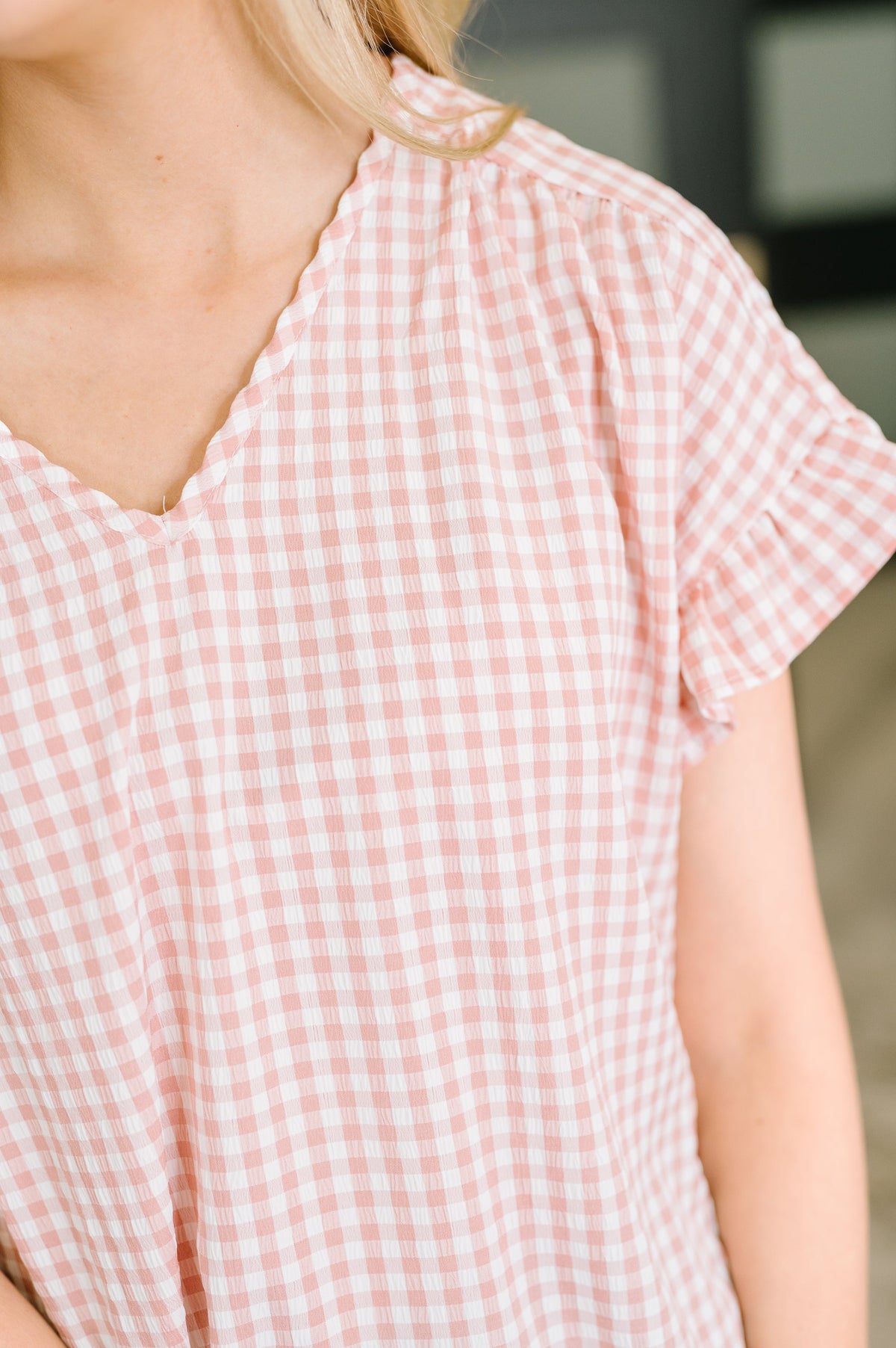 Close-up of a person wearing a light pink and white gingham short-sleeve top with a ruffled scalloped V-neckline.