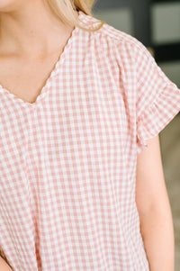 Close-up of a person wearing a light pink and white gingham short-sleeve top with a ruffled scalloped V-neckline.