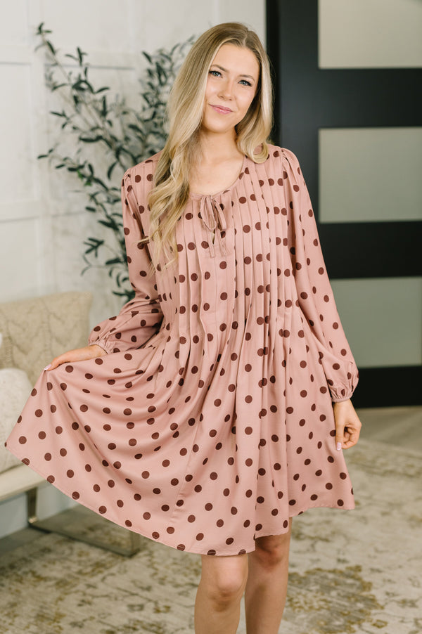 A woman modeling a long-sleeve, knee-length dress with a dusty pink base and dark brown polka dots, standing in an indoor setting.
