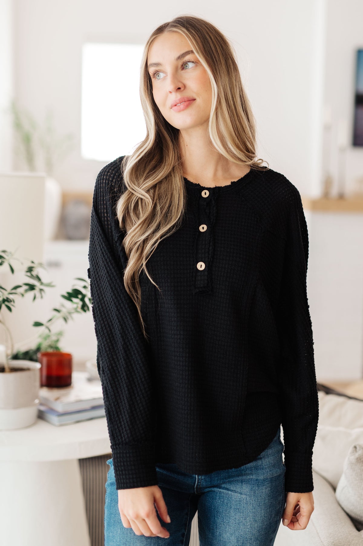 A woman wearing a black textured long-sleeve henley top with gold buttons, paired with blue jeans, standing in a bright, modern room.