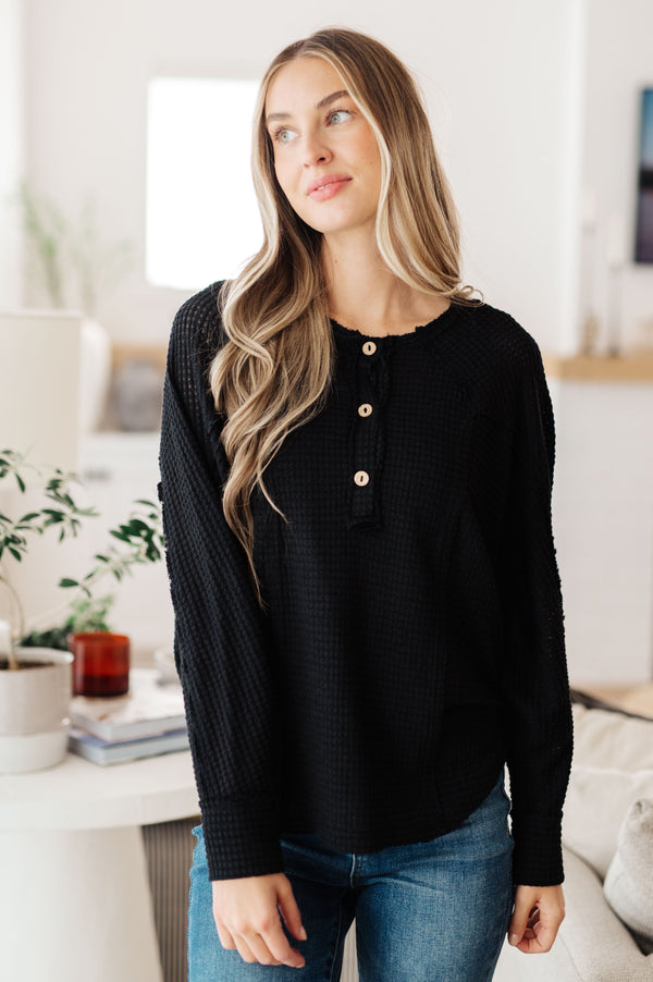 A woman wearing a black textured long-sleeve henley top with gold buttons, paired with blue jeans, standing in a bright, modern room.