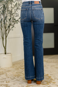A person shown from the back wearing blue denim flare jeans with brown block heels, standing on a patterned rug in an indoor setting.