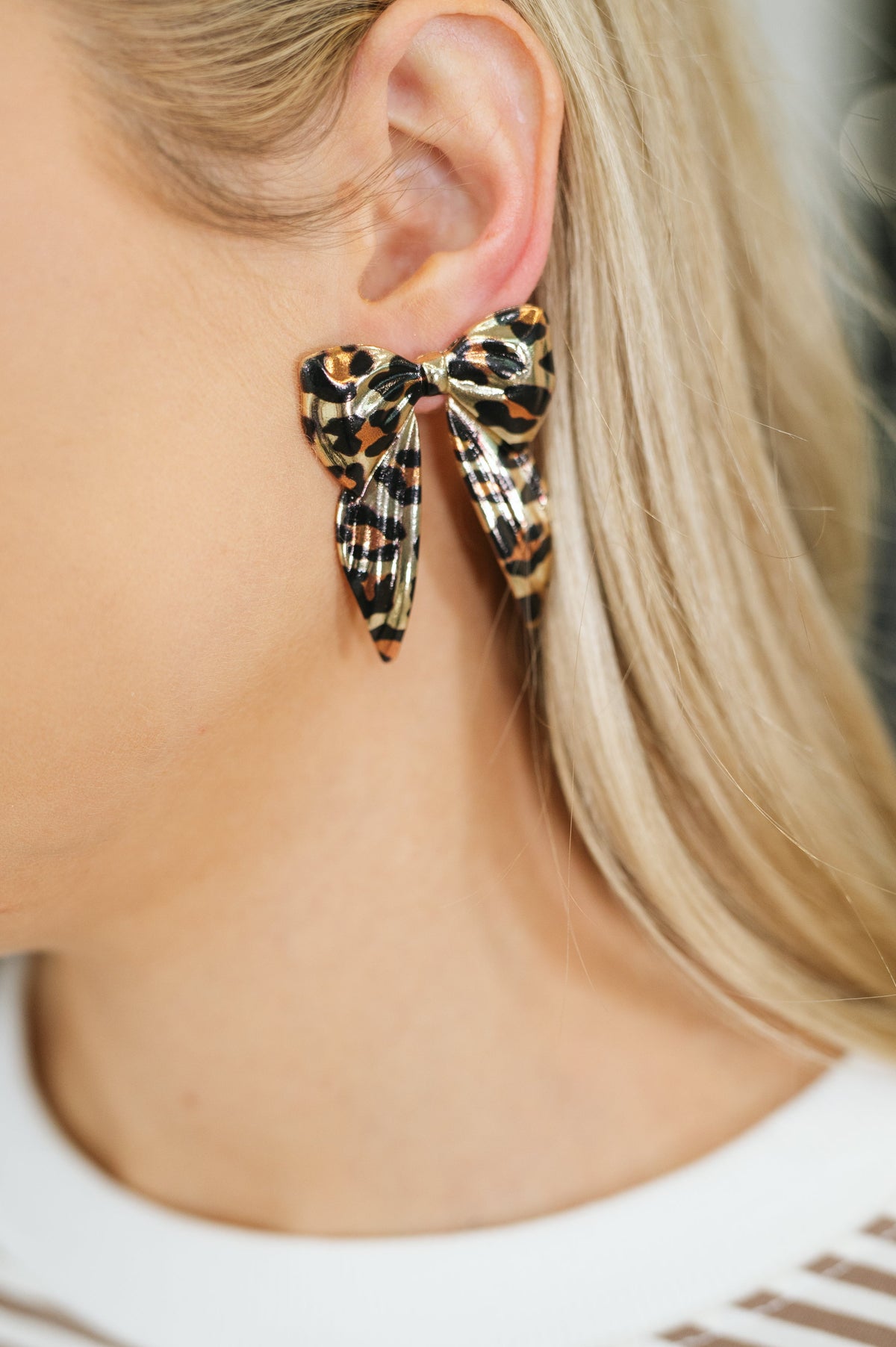 A close-up of a model's ear wearing a leopard-print bow stud earring. The earring features a black, orange, and beige animal print pattern.