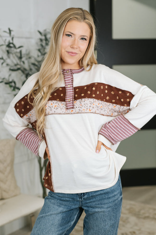 A woman modeling a long-sleeve patchwork top with a white base, brown patterned panels, and striped accents, paired with blue jeans in an indoor setting.