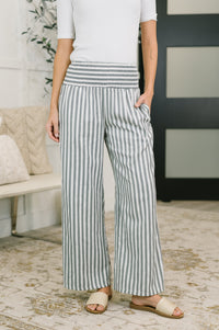 A person wearing high-waisted, wide-leg pants with vertical blue and white stripes, paired with a white top and beige slide sandals in a home interior setting.