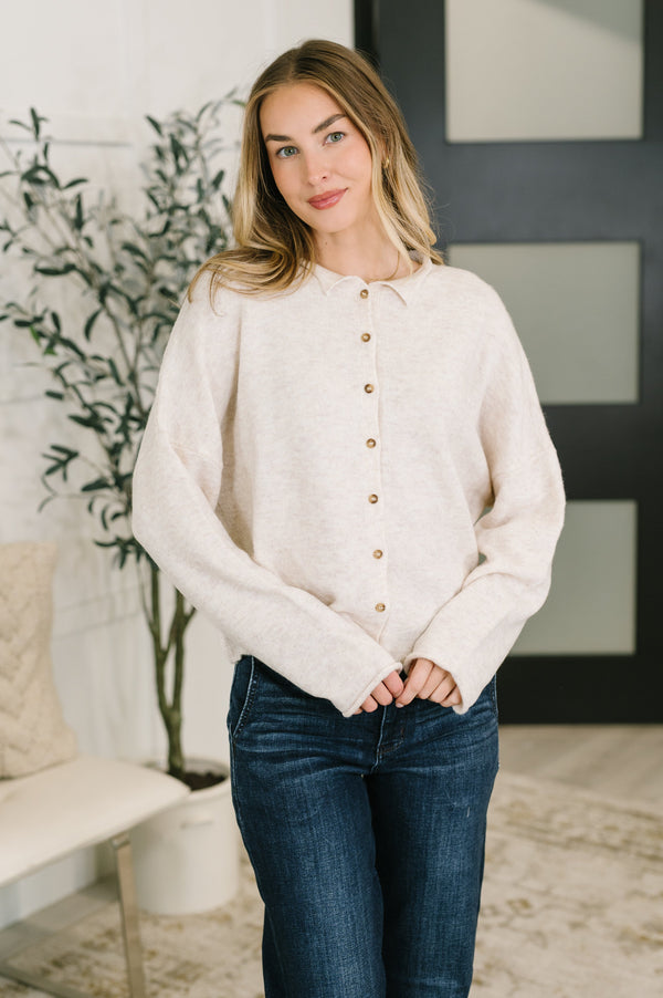 A woman wearing a cream-colored, long-sleeve knit button-up sweater and dark blue jeans, standing in a modern indoor setting with a plant and a bench in the background.