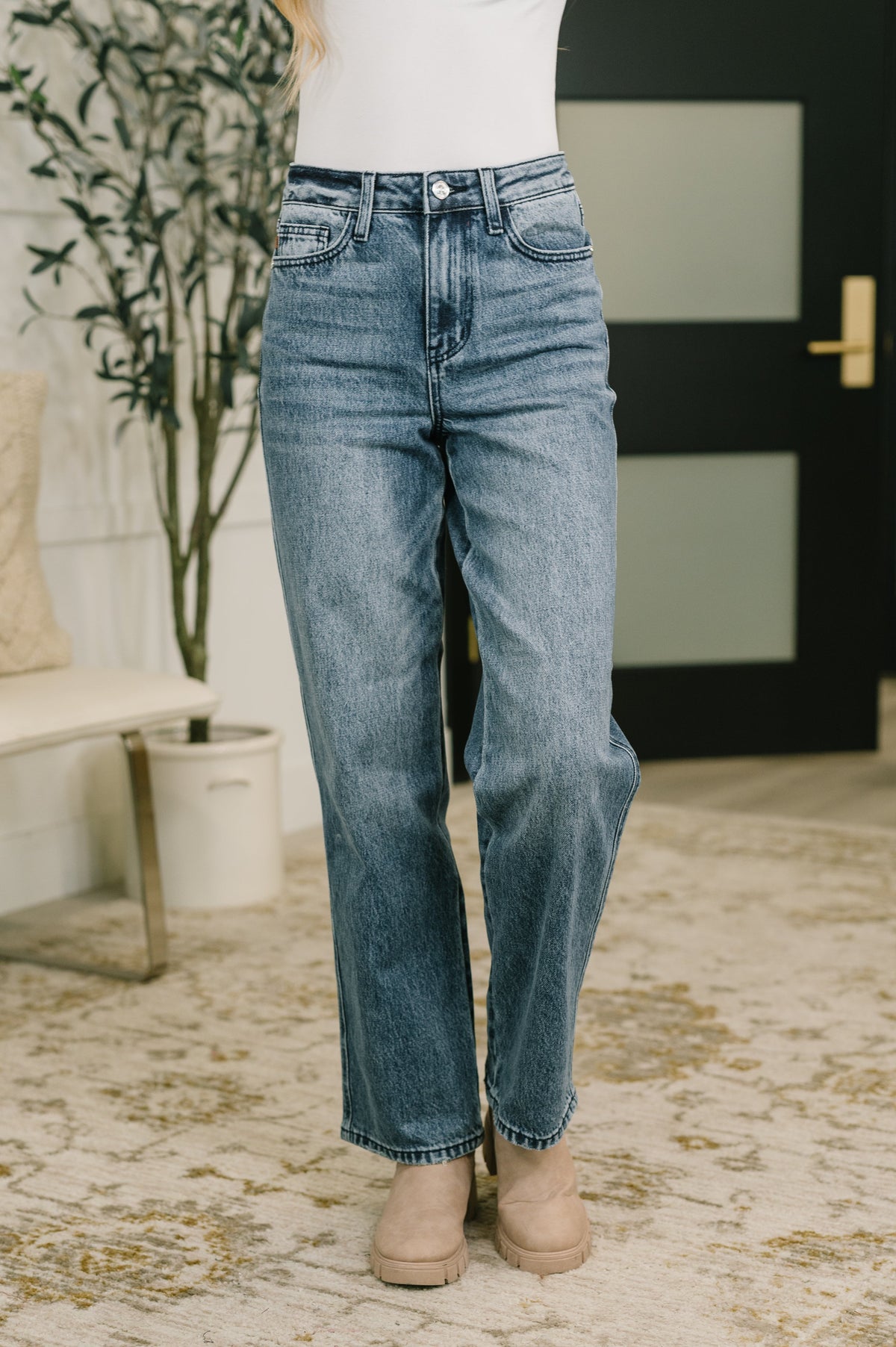 A person wearing light-wash blue straight-leg jeans, a white top, and beige ankle boots, standing on a patterned rug in an indoor room with a plant and dark door in the background.