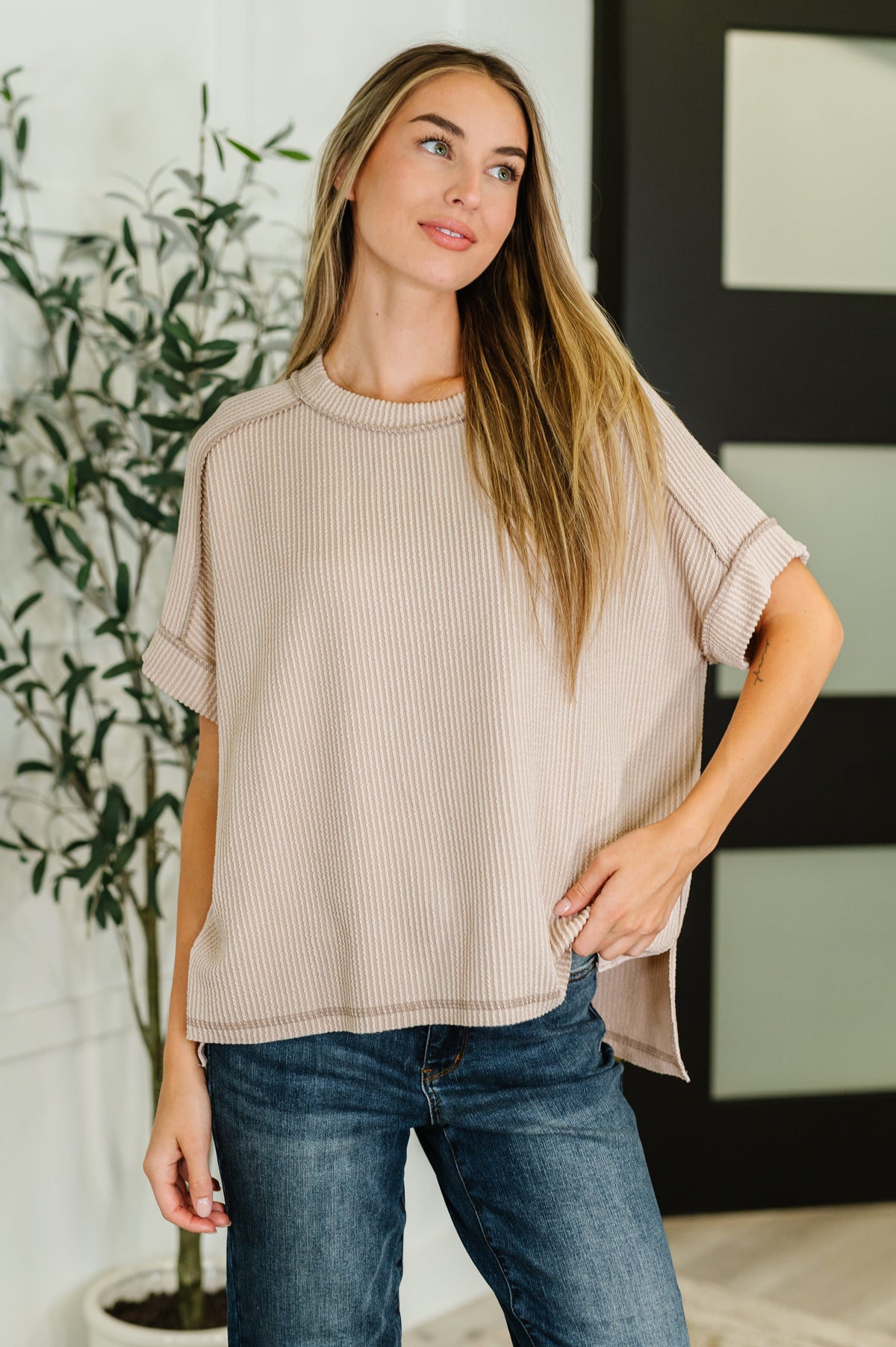 Woman wearing a beige top and blue jeans standing indoors with a plant in the background