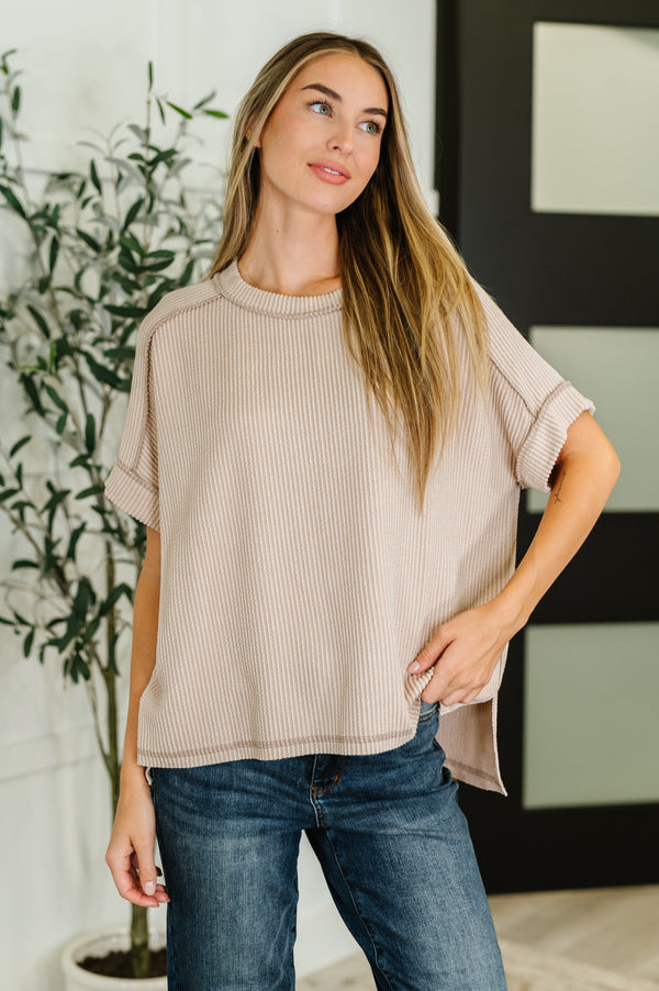 Woman wearing a beige top and blue jeans standing indoors with a plant in the background