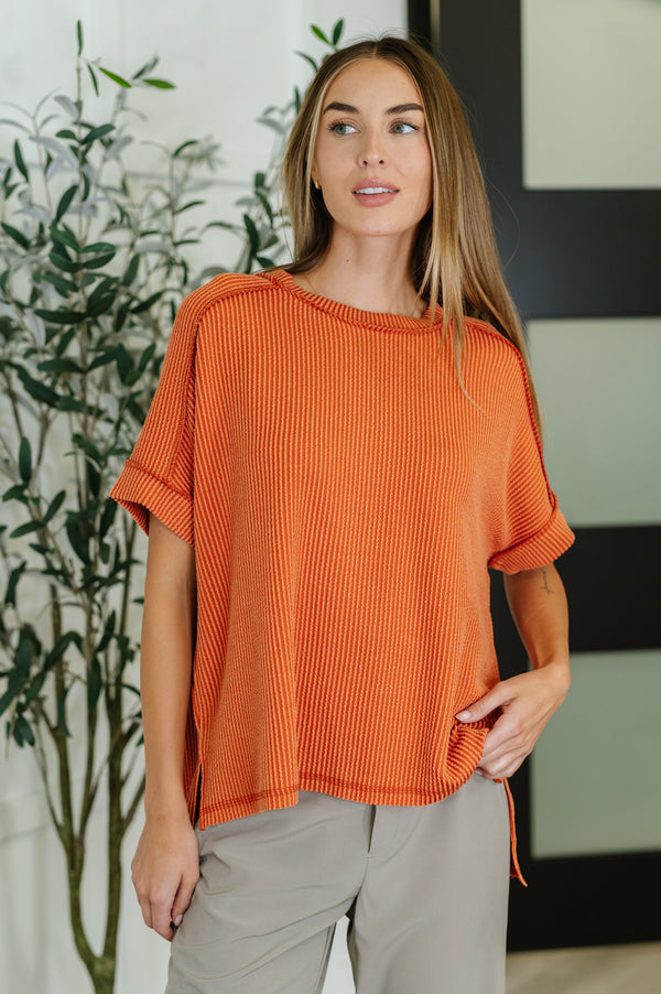 Woman wearing an orange ribbed top and beige pants standing indoors with a plant in the background