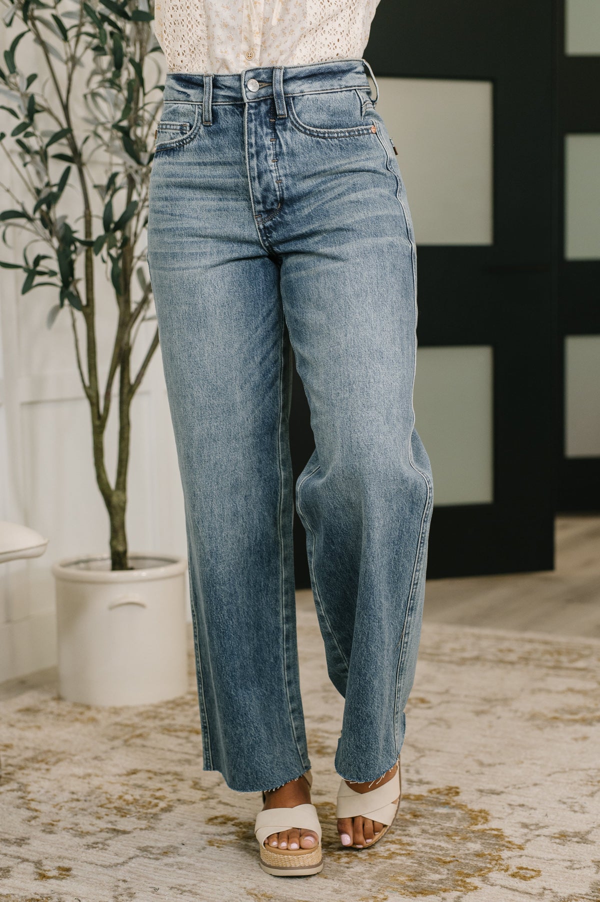 Person wearing blue jeans standing indoors with a plant and decorative wall in the background