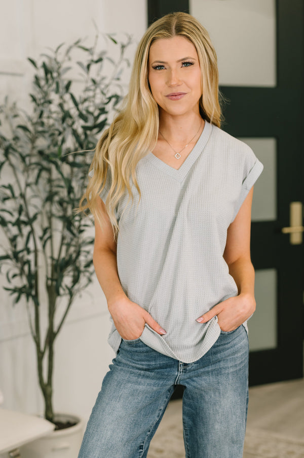 Woman wearing a light gray sleeveless top and blue jeans standing indoors.