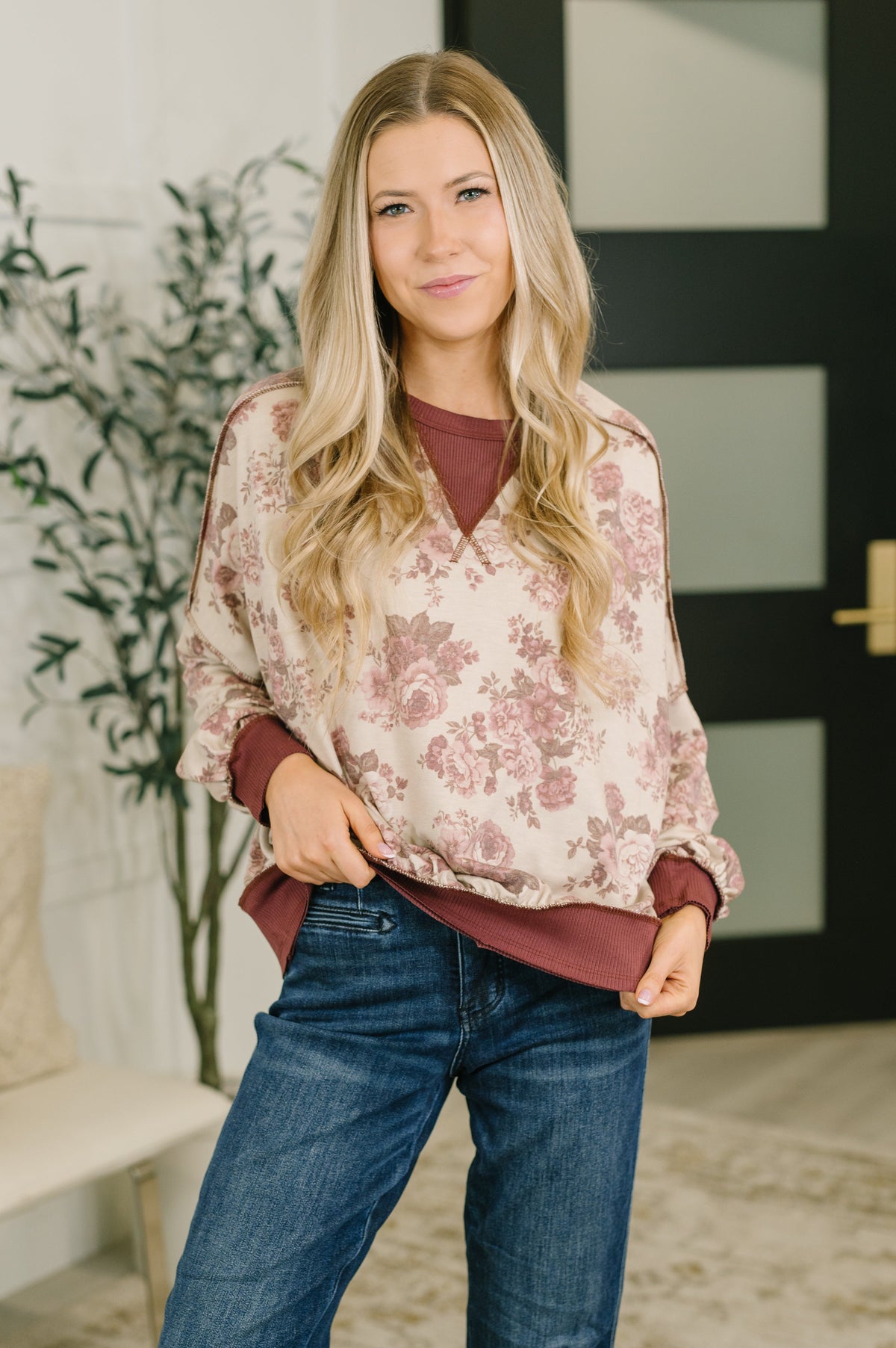 A woman modeling a long-sleeve V-neck blouse with a dusty rose floral pattern on a cream background, layered over a dark burgundy top and paired with blue jeans.