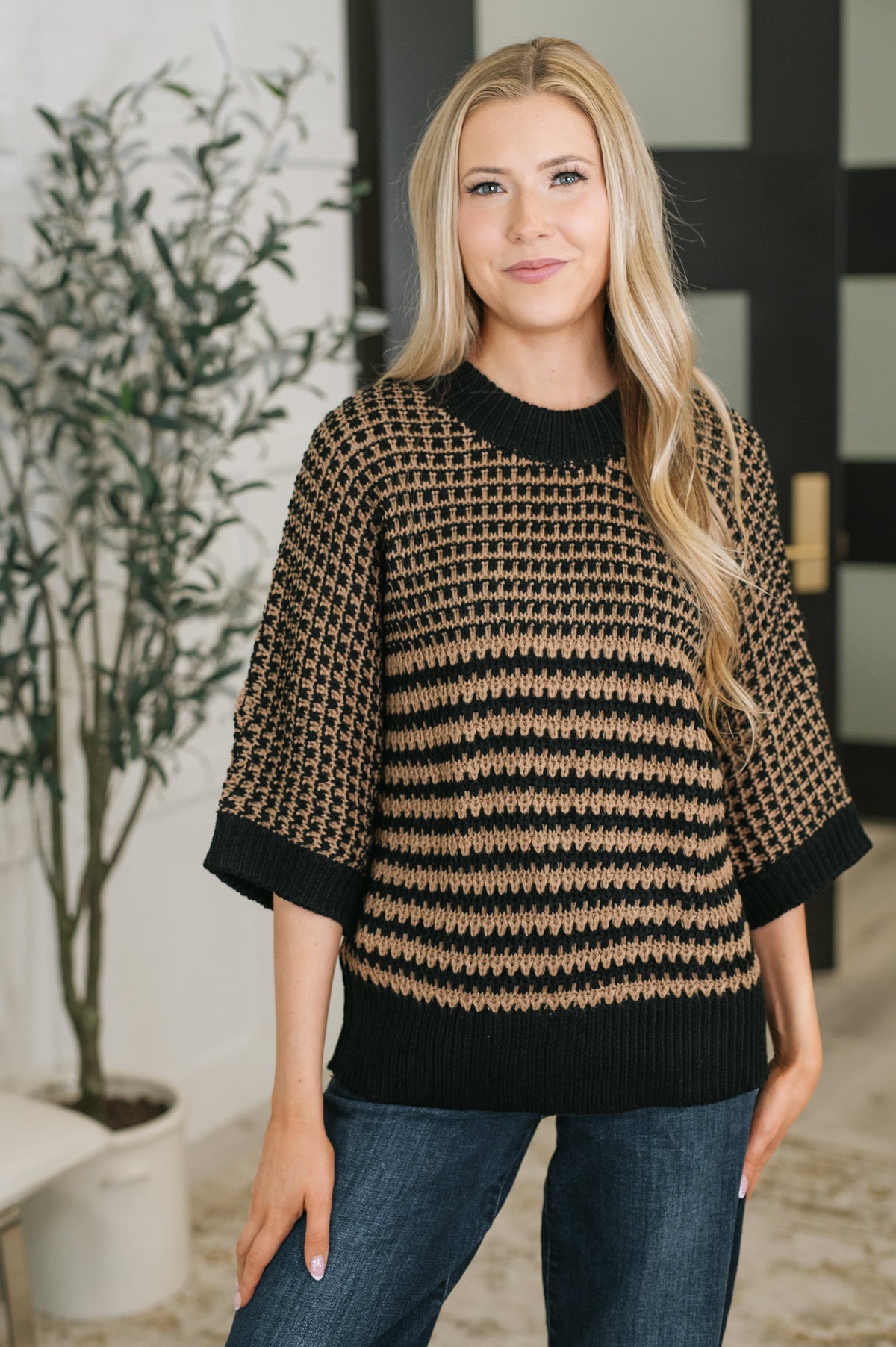 A woman wearing a black and tan patterned knit sweater with three-quarter sleeves and a crew neck, paired with blue jeans, standing in an indoor setting.