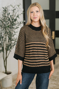 A woman wearing a black and tan patterned knit sweater with three-quarter sleeves and a crew neck, paired with blue jeans, standing in an indoor setting.