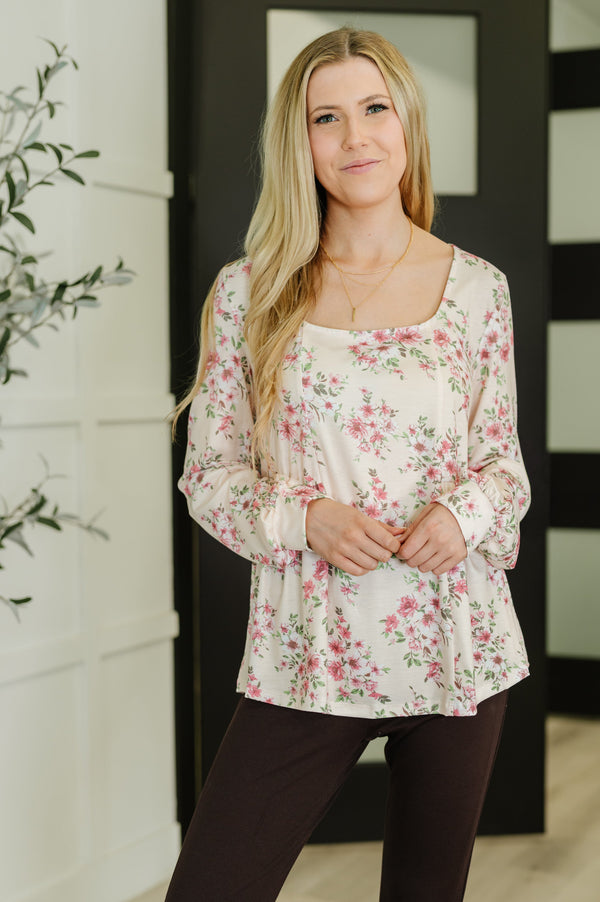 A woman modeling a long-sleeve scoop-neck blouse with a pink and green floral print on a cream background, paired with dark brown pants.