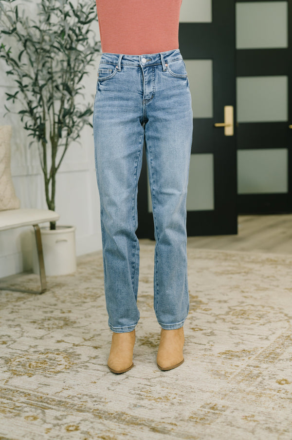 A person wearing light-wash blue jeans, a coral pink top, and tan ankle boots, standing indoors on a patterned rug in front of a black door.